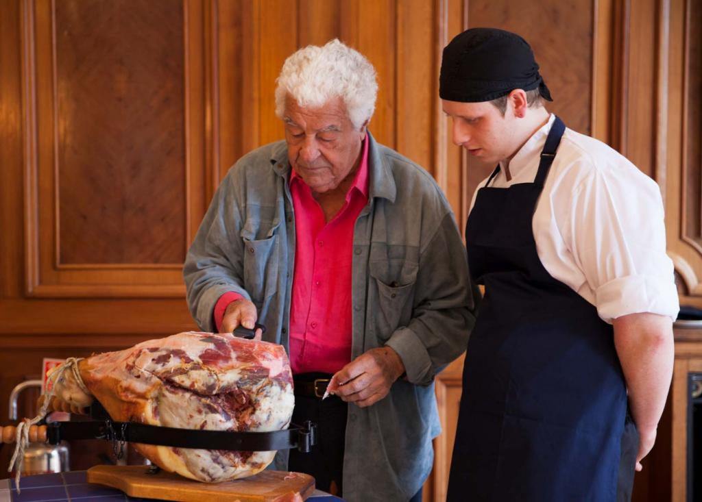 Antonio Carluccio using a knife one in his right hand to slice some ham from a cured ham leg, watched closely by a younger chef